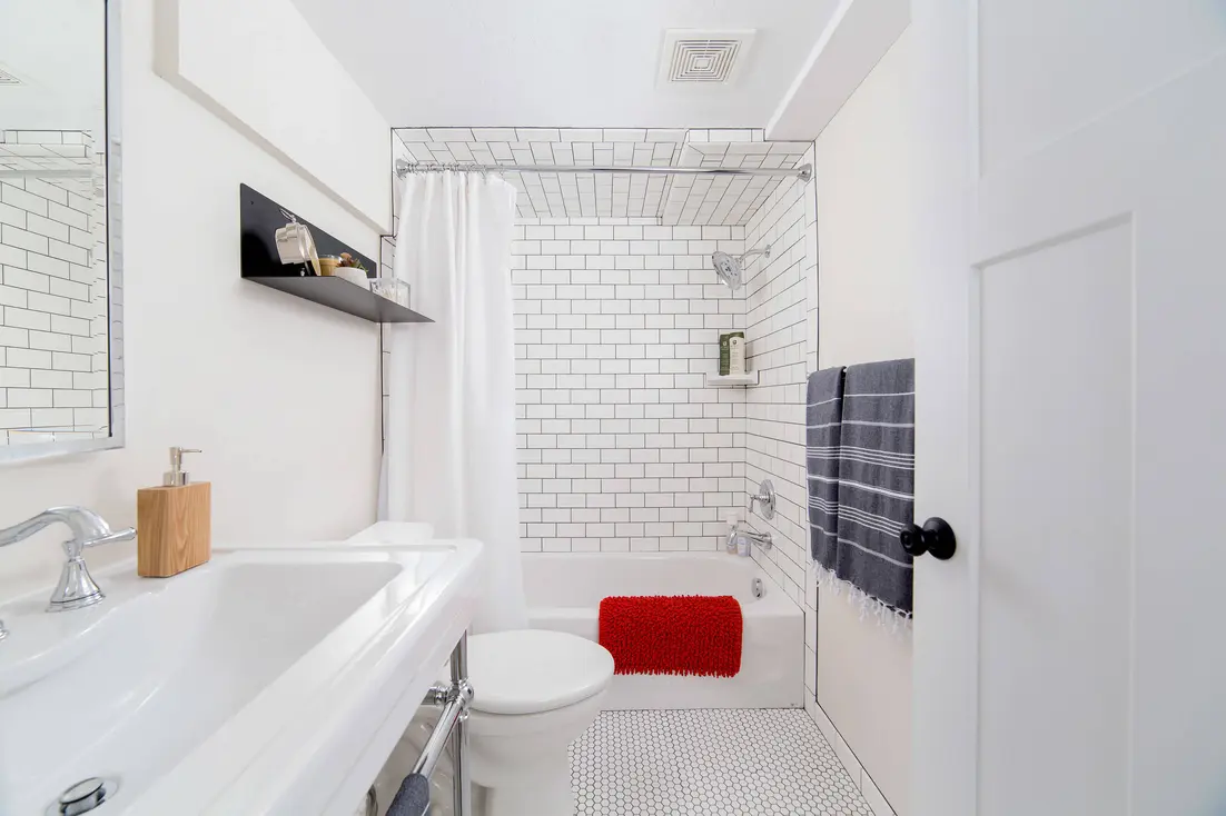 Modern bathroom with a white tiled shower, bathtub, and a red bath mat on the floor.