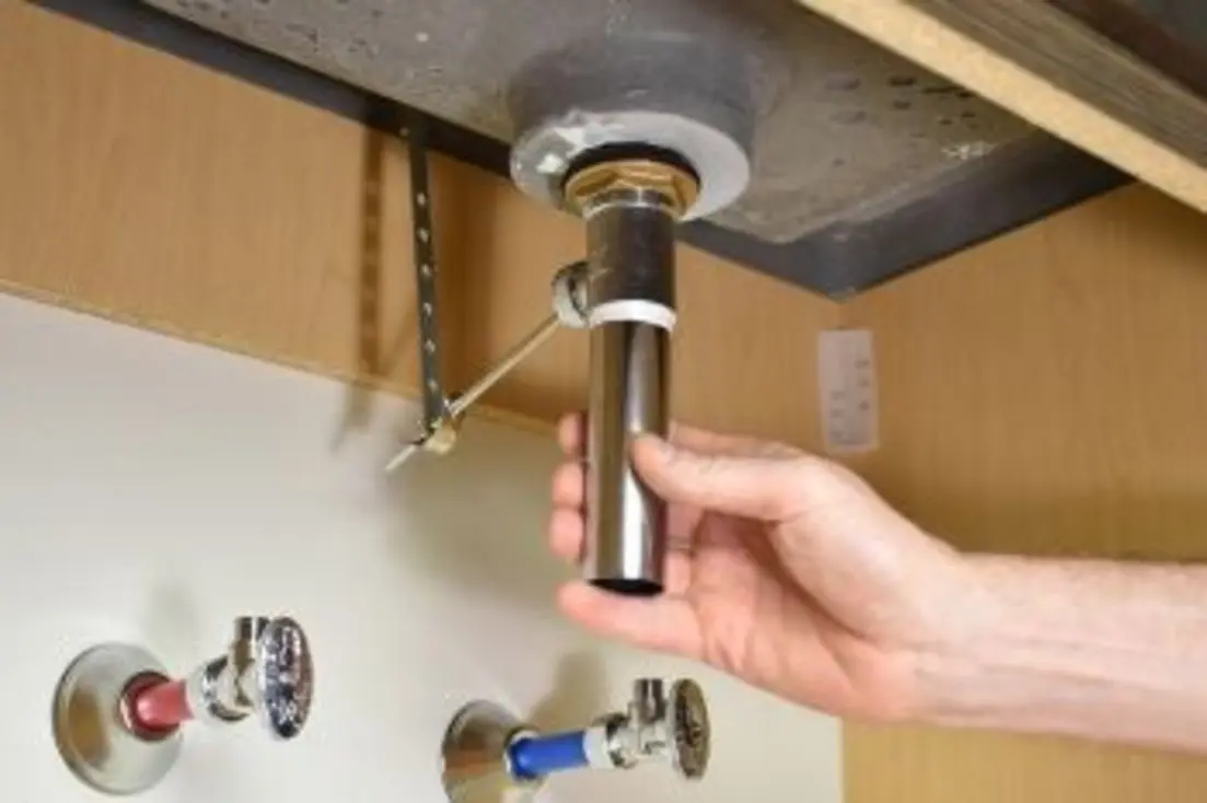 A person’s hand adjusting a plumbing pipe under a bathroom sink.