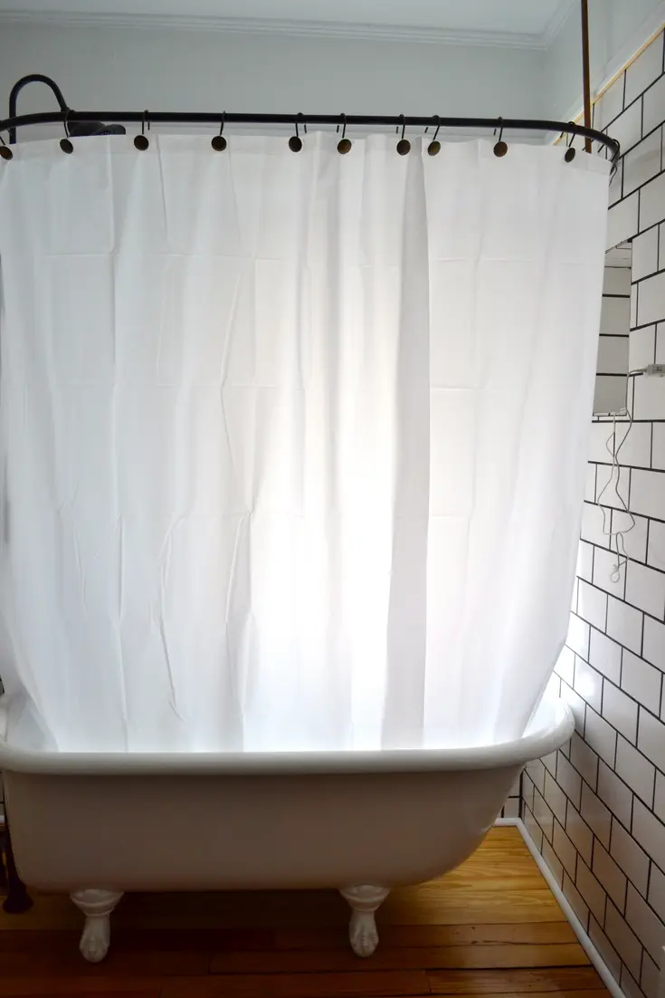 A white shower curtain hangs above a clawfoot bathtub in a tiled bathroom.
