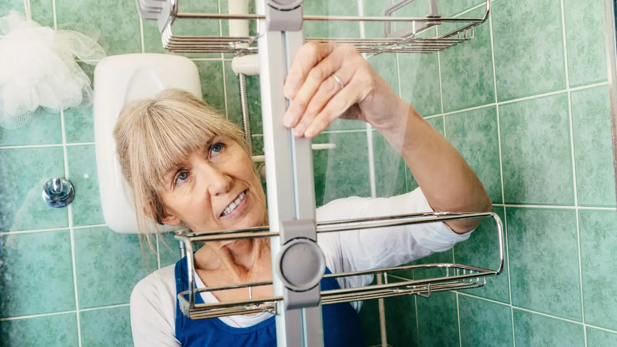 An older woman adjusts a metal shower caddy in a tiled bathroom.