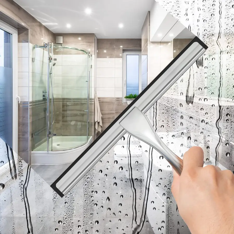 A hand holding a squeegee clears water from a glass shower door in a bright bathroom.