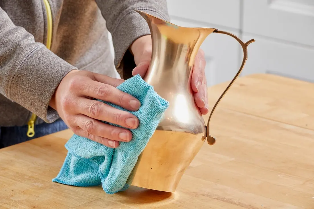Person cleaning a brass pitcher with a blue cloth on a wooden surface.