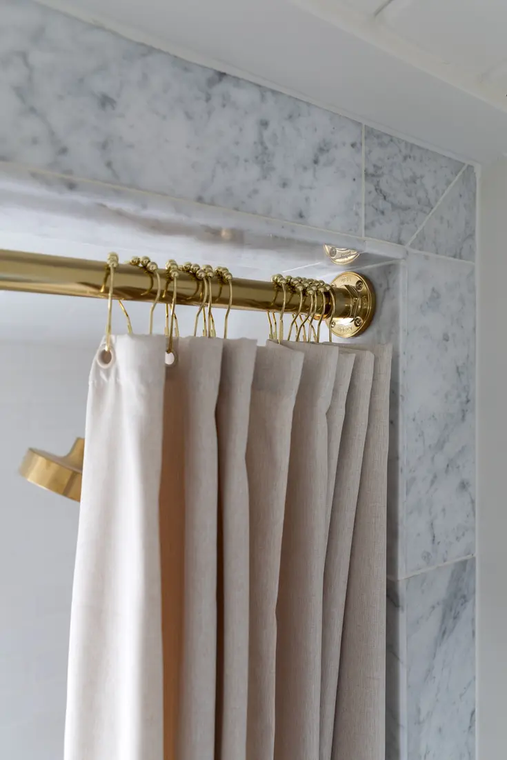 Shower curtain rod with beige curtains and gold hardware against marble wall.