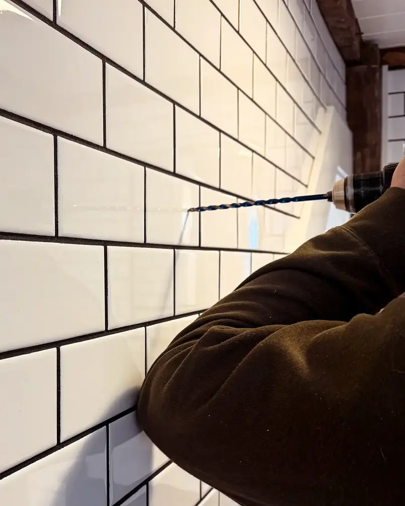 A person drilling into a tiled wall with a handheld drill and blue drill bit.