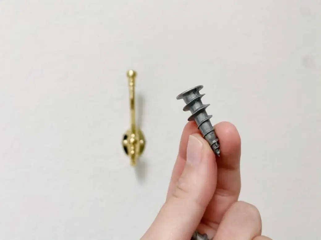 Hand holding a metal screw near a gold towel hook on a wall.