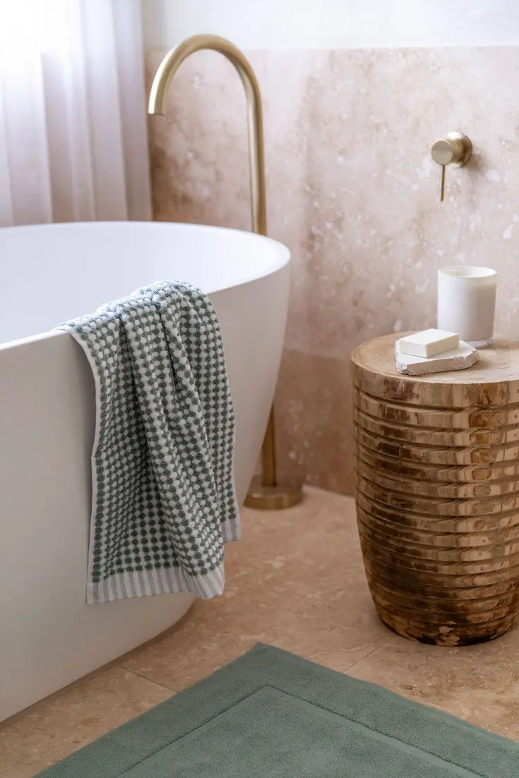 A textured green bath mat, striped towel, and decorative wooden stool near a freestanding bathtub.