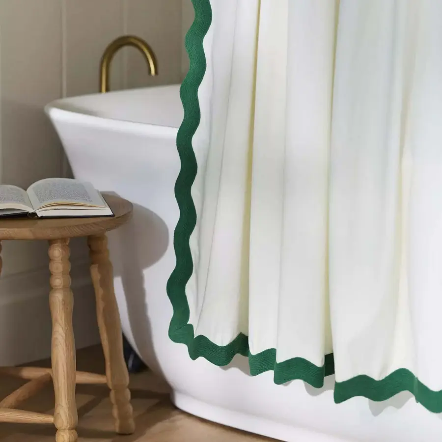 A white shower curtain with green edging near a freestanding tub and a wooden stool with a book.