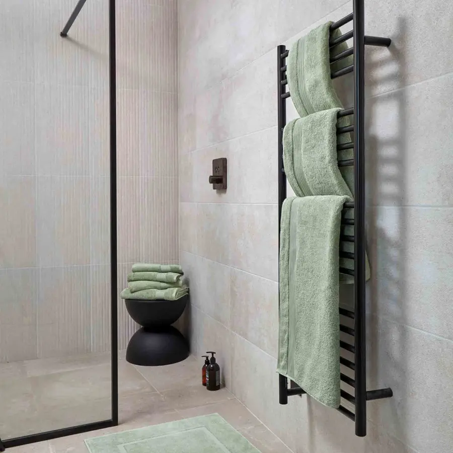 A bathroom with a black towel rack holding neatly folded green towels against a light tiled wall.