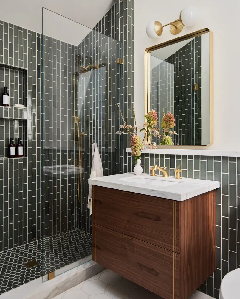 Bathroom with a glass shower, dark subway tile, wooden vanity, and gold fixtures.